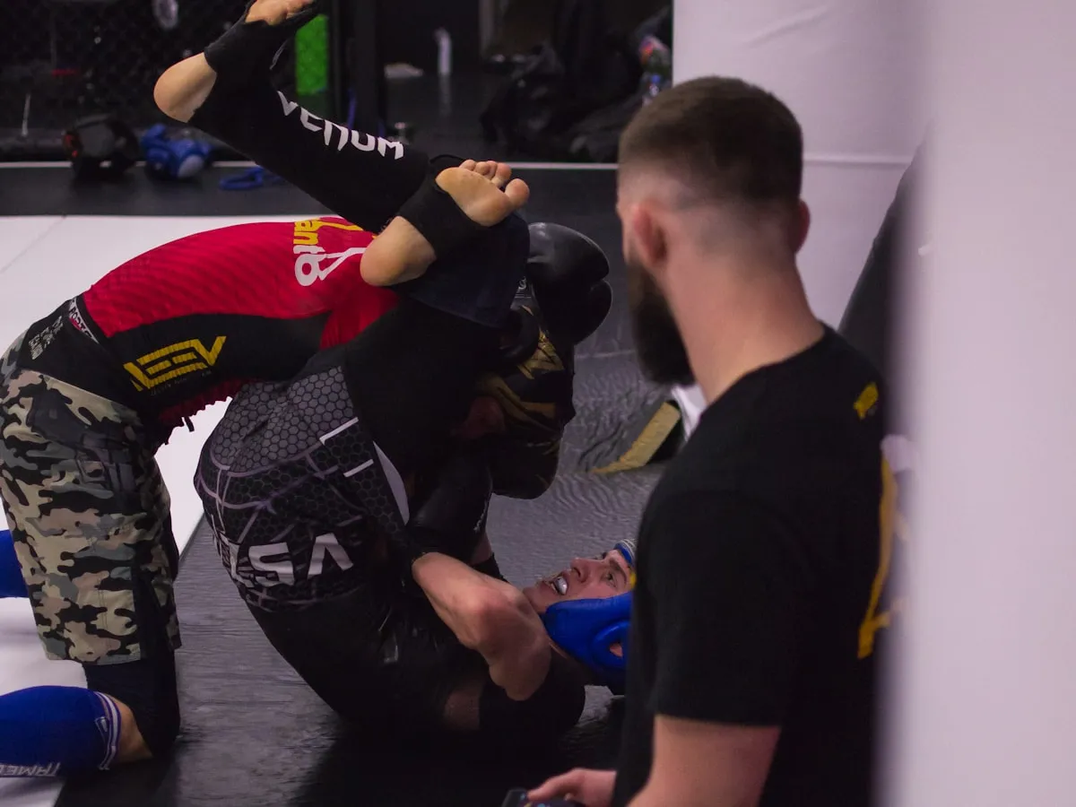 Two athletes sparring no-gi jiu jitsu on the gym mat as a coach watches, an MMA cage in the background